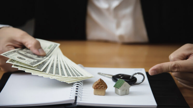 A close-up, realistic photo of a person in a business suit sitting at a wooden desk. One hand holds a fanned-out stack of US hundred-dollar bills, while the other hand points toward a notebook. On the open notebook, there are two small model houses and a car key, symbolizing financial planning, real estate, and asset management.
