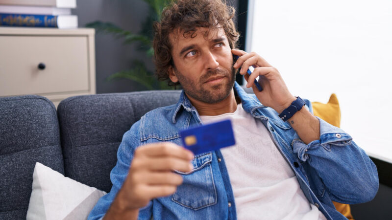 Young hispanic man talking on smartphone holding credit card sitting on sofa at home