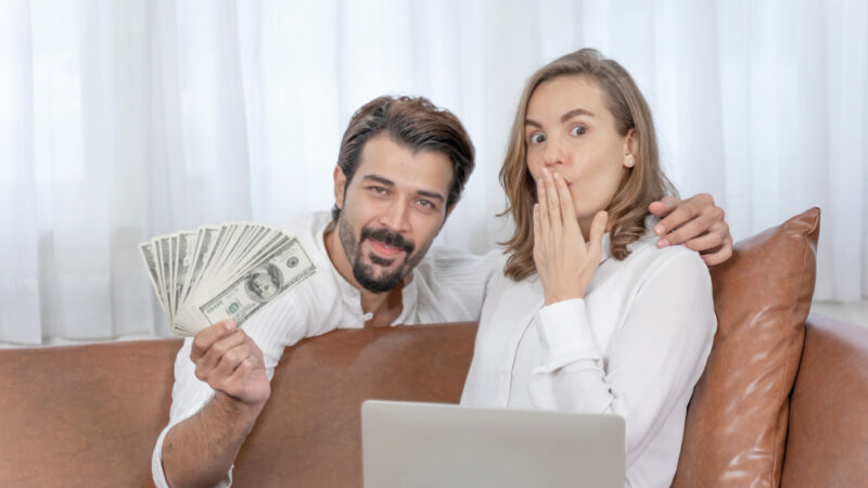 A medium shot of a young couple sitting on a brown leather sofa in a bright room. The man has his arm around the woman and holds a fanned-out stack of hundred-dollar bills, smiling confidently at the camera. The woman looks surprised, with her hand over her mouth, while a laptop sits open in front of them. The image illustrates a successful conversation about finances, budgeting, or shared savings goals.