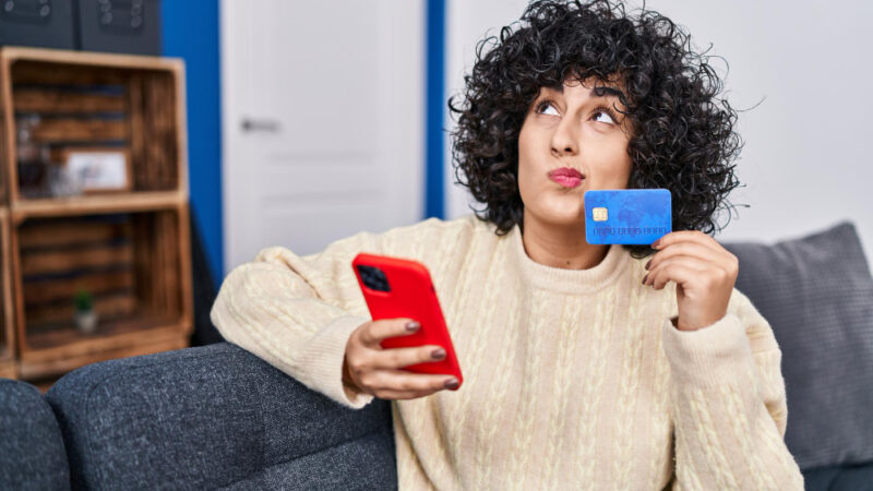 Young middle east woman using smartphone and credit card sitting on sofa at home