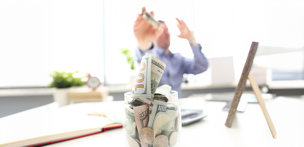 Rich man throwing us banknotes in big jar full of money at workplace
