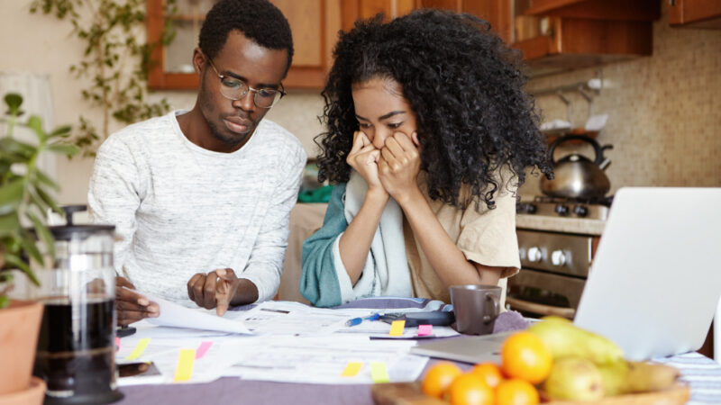 Stressed young african wife holding hands on her face, listening in desperation to her husband reading notification, informing that they have to move out of their aparment because of non-payment