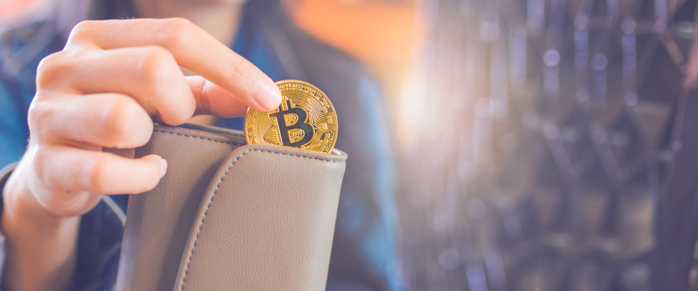 The hand of a woman is pulling a bitcoin coin out of a wallet.
