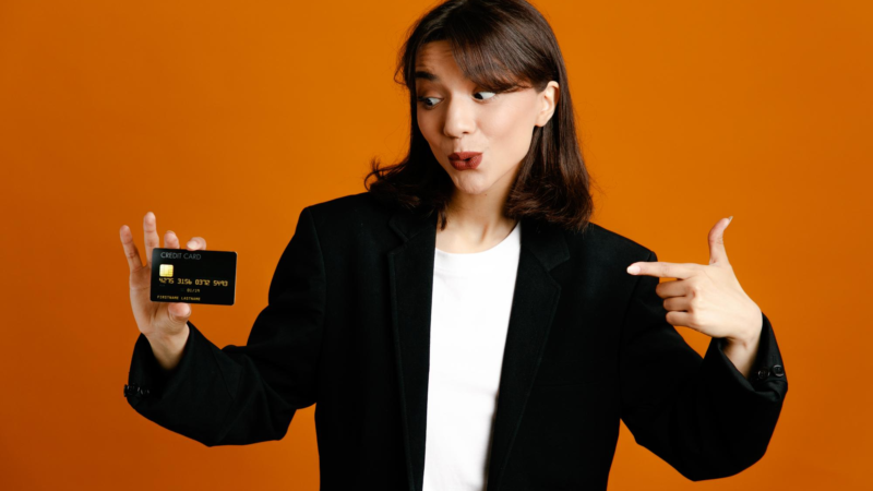 Impressed holding card young beautiful female wearing black jacket isolated on orange background