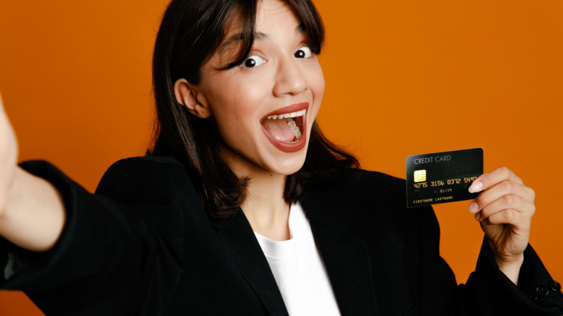 Surprised holding camera with card young beautiful female wearing black jacket isolated on orange background